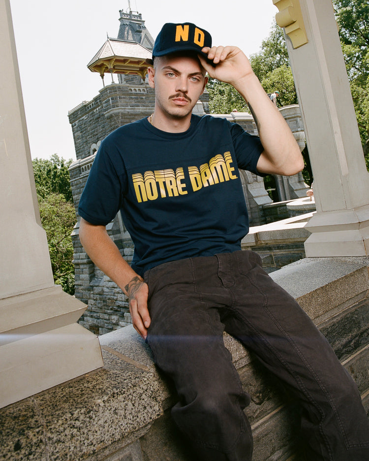 A man sits on a stone ledge outdoors, wearing the Ebbets Field University of Notre Dame Short Sleeve Football Tee, dark pants, and a dark cap with yellow ND letters. He holds his cap’s brim and looks at the camera amid trees and stone structures.