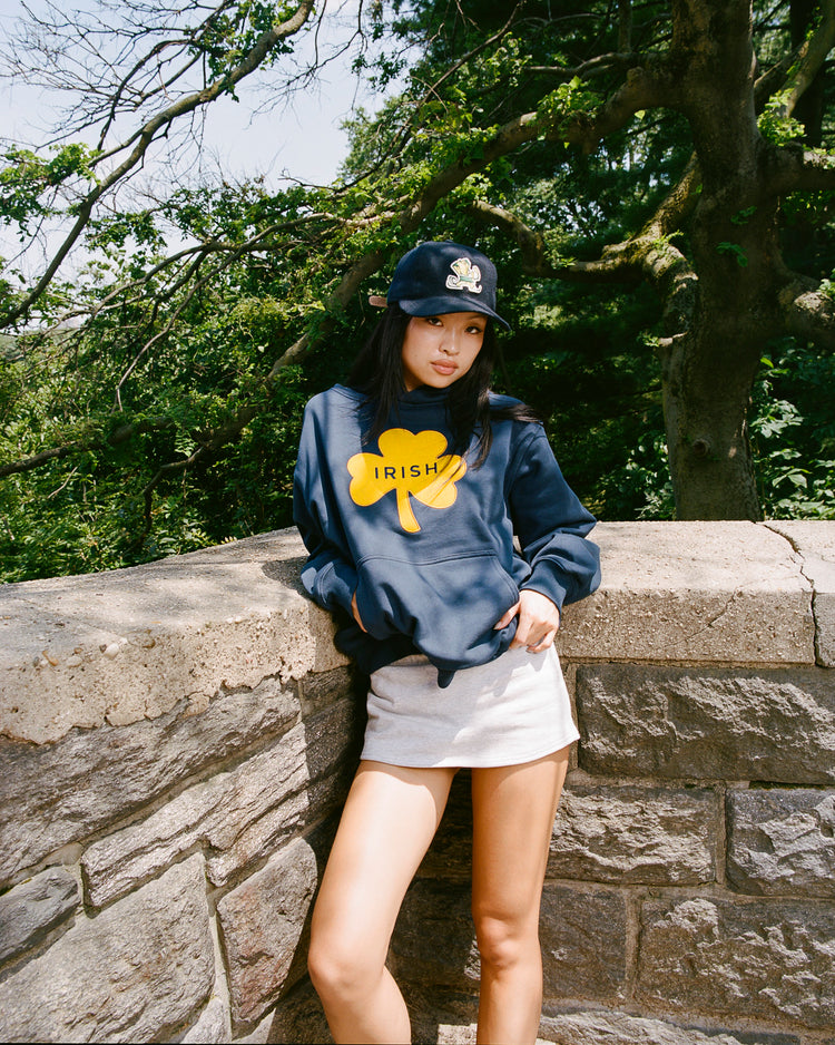 A young woman in a navy hoodie with a yellow shamrock and IRISH stands by a stone wall outdoors, wearing the EFF University of Notre Dame Leprechaun Authentic Wool Ballcap, plus a light skirt, with trees and sunlight in the background.