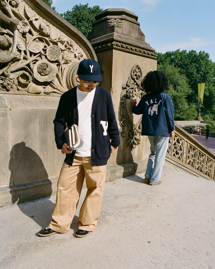 Two people stand by an ornate stone staircase outdoors. One, holding books, wears a Y cap and cardigan; the other, in light jeans, sports the Ebbets Field Yale University Cotton Grounds Crew Jacket. Lush green trees are behind them.