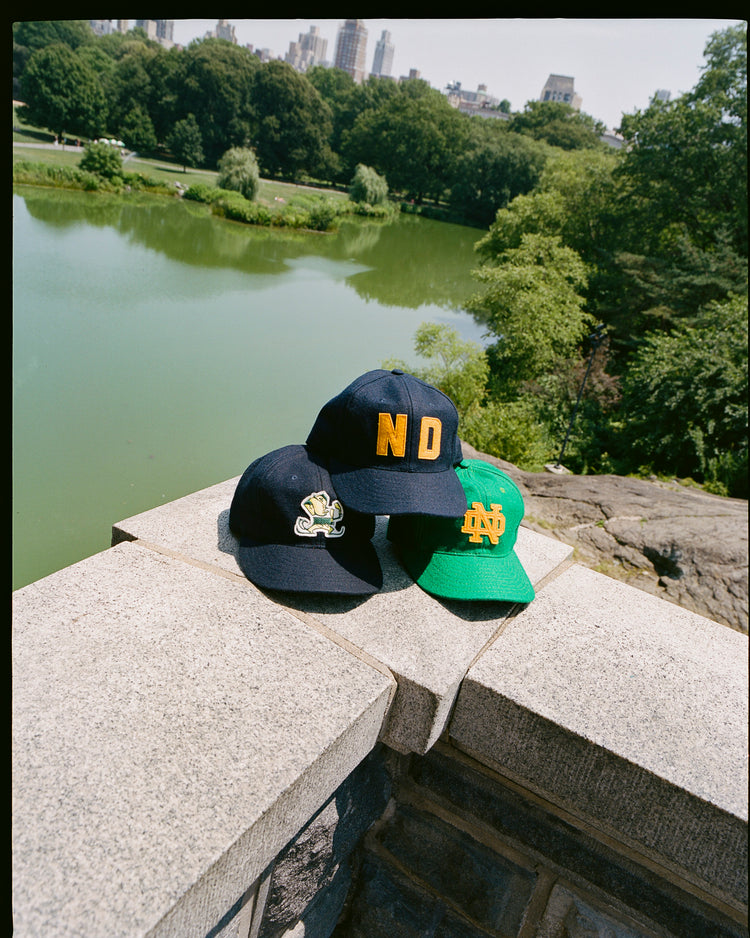 Three EFF University of Notre Dame ND Authentic Wool Ballcaps—two navy and one green—sit on a stone ledge by a pond with trees and a city skyline in the background, capturing Notre Dame’s rich heritage.