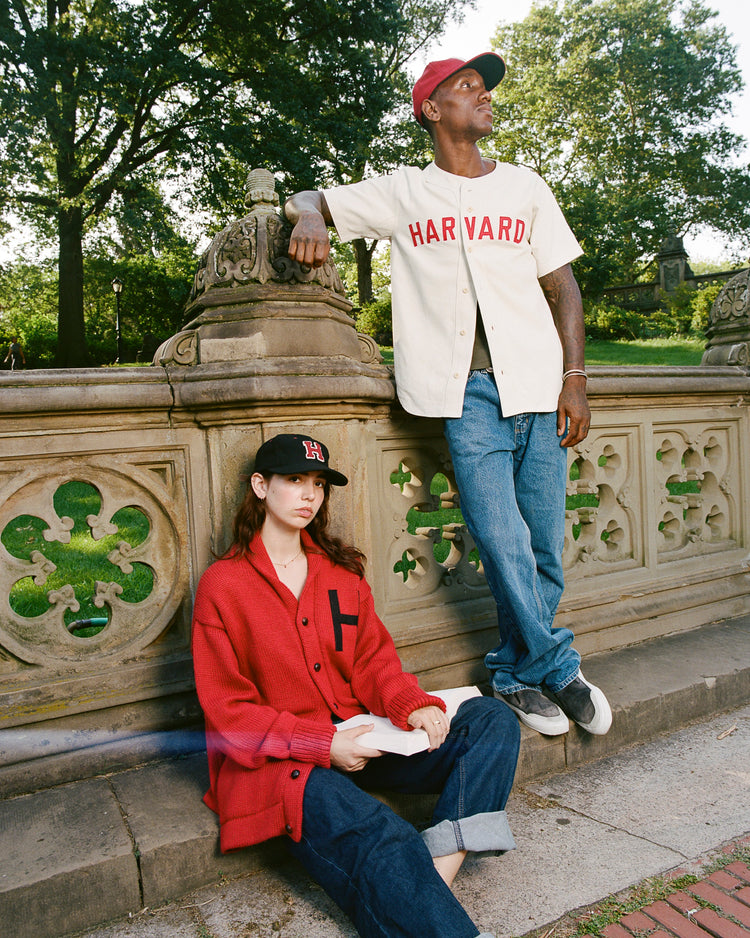 A woman in a red sweater and cap sits on a sidewalk holding a book, while a man wearing the Ebbets Field Harvard University Cotton Twill Baseball Jersey and jeans stands beside her, leaning on a stone railing in a park with green trees.