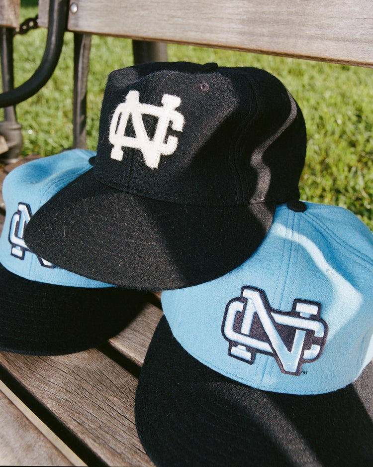 Three baseball caps, including the Ebbets Field Flannels University of North Carolina 1960 Vintage Ballcap, rest on a wooden bench—one black with white logo and two light blue with black-and-white logos—against lush green grass.