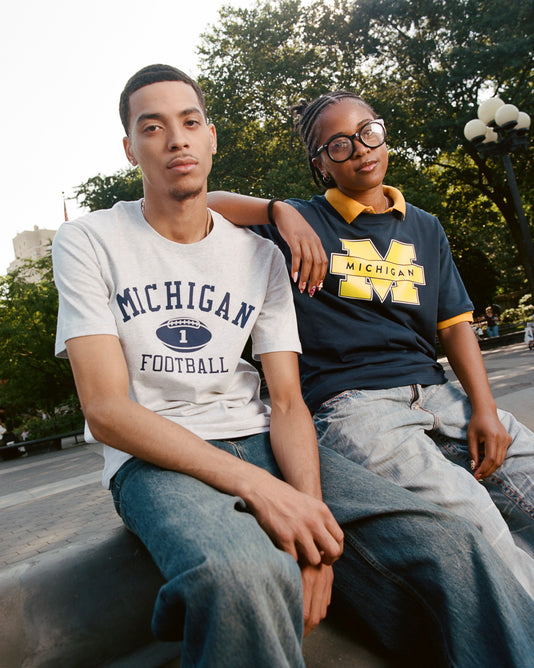 Two people sit outdoors on a concrete ledge wearing Ebbets Field’s MICHIGAN SS TEE H-GRY and jeans. The person on the left wears this heather grey Michigan tee, while the other wears a navy Michigan shirt with large glasses.