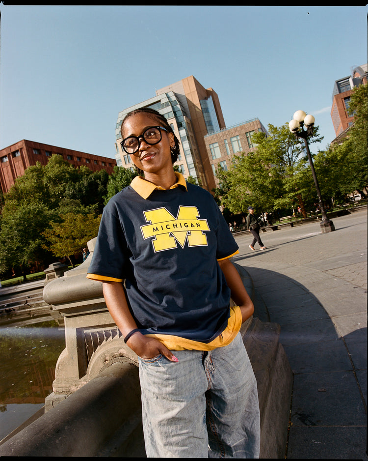 A person wearing Ebbets Field’s MICHIGAN SS TEE NAVY stands by a fountain in an urban park, surrounded by trees and modern buildings under a clear sky.
