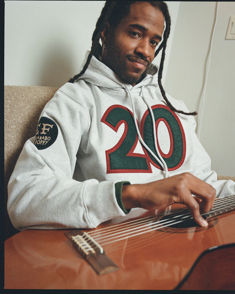 A man with dreadlocks wears an Ebbets Field Karabo Poppy x Ebbets Field Josh Gibson Heavyweight Cotton Pullover Hoodie featuring a bold 20 on the front as he sits on a couch, smiling softly while playing an acoustic guitar.