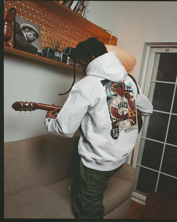 A person with long braids wearing an Ebbets Field Karabo Poppy x Josh Gibson Heavyweight Cotton Pullover Hoodie and dark pants stands on a sofa playing guitar. Shelves with books, a lamp, and framed photos evoke the spirit of the Negro Leagues.