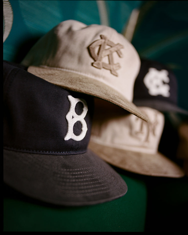 A close-up of three Ebbets Field ballcaps, featuring the Brooklyn Royal Giants 1925 Cotton Twill Suede Ballcap in navy and two beige Negro League caps, all with embroidered logos, arranged in a row against a green background.