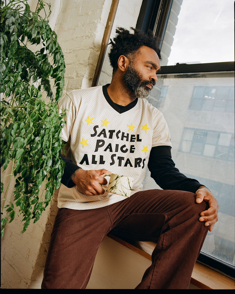A bearded man sits on a windowsill with a mug, wearing the Ebbets Field Satchel Paige All-Stars Mesh V-Neck Jersey over a long-sleeve shirt and brown pants beside a leafy plant and large window—honoring Negro League baseball history.
