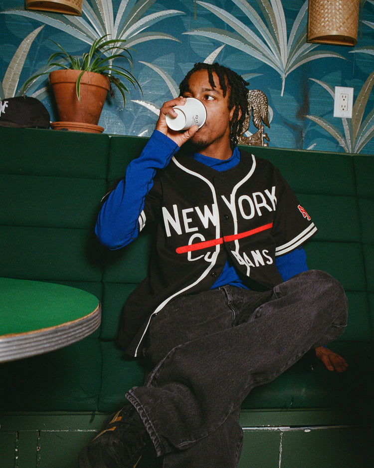 A person sits on a green bench, sipping from a white cup. They wear an Ebbets Field New York Cubans Cotton Twill Baseball Jersey over a blue shirt and black pants. Plants and patterned wallpaper complete the charming scene.