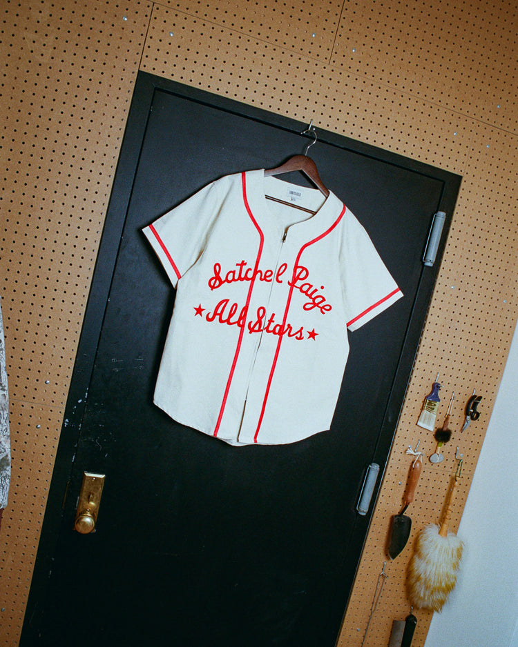 An Ebbets Field Satchel Paige All-Stars 1933 Cotton Twill Jersey with red trim hangs on a black door, framed by pegboard walls and cleaning brushes on hooks.