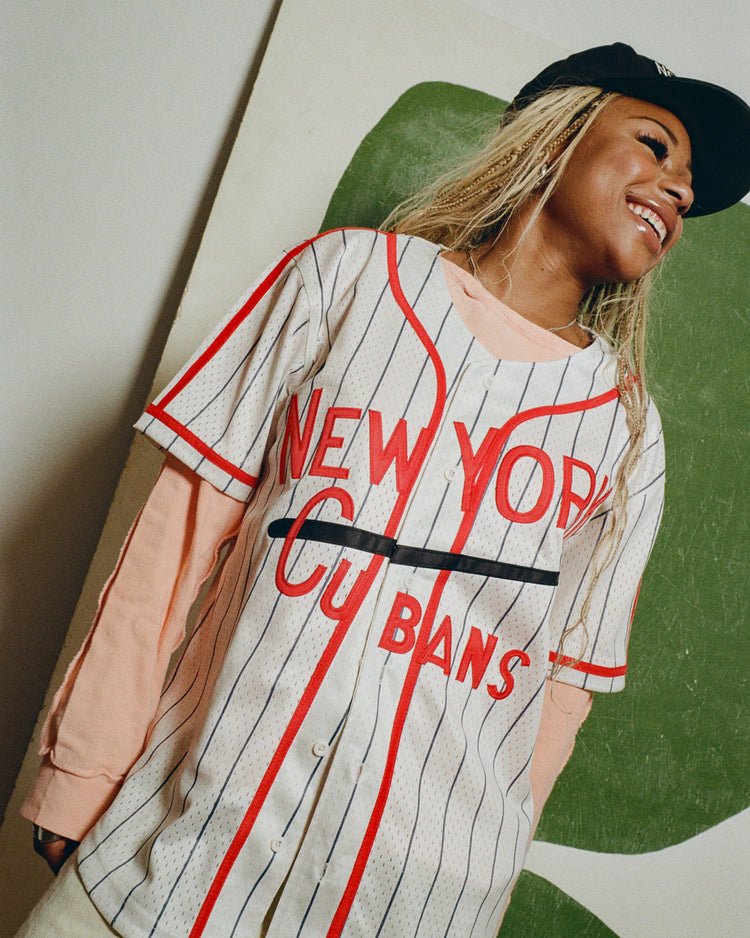 A smiling person in a black cap and layered shirts, including an Ebbets Field New York Cubans Mesh Button-Up Jersey, stands before an abstract green and white background.