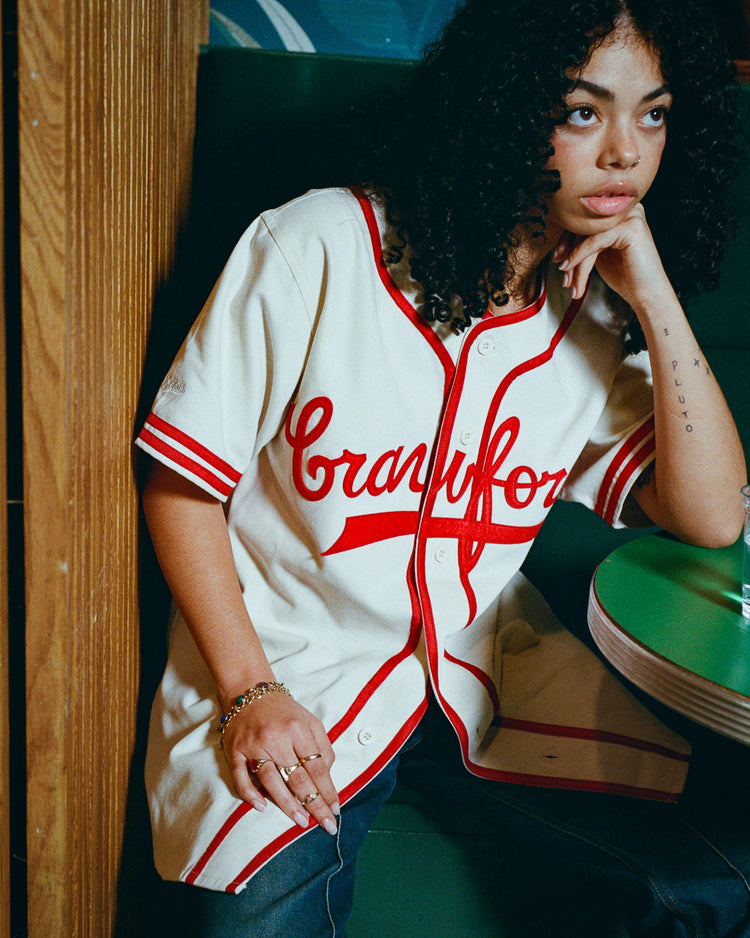 A person with curly hair wears an Ebbets Field Pittsburgh Crawfords 1933 Cotton Twill Jersey in cream and red, sitting in a booth and gazing thoughtfully to the side, reflecting on Black baseball heritage.