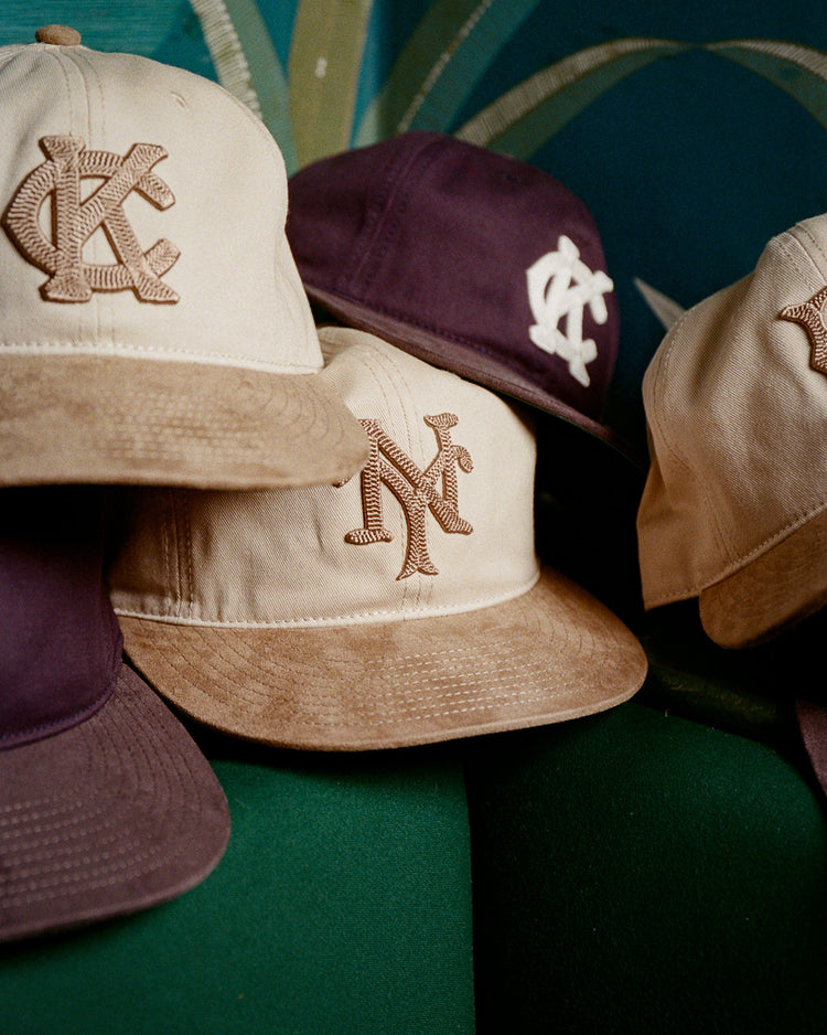 The Ebbets Field New York Cubans 1948 Cotton Twill Suede Ballcap, in beige and dark purple with embroidered front letters, is displayed on a green surface against a blue patterned background.