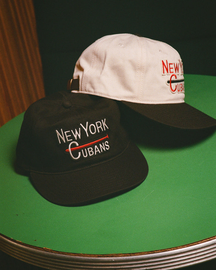 Two New York Cubans Cotton Twill Ballcaps by Ebbets Field—one black, one white—sit on a green table, each featuring 