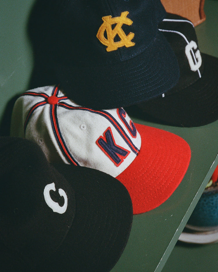 Four baseball caps, including the Ebbets Field Kansas City Monarchs 1945 Authentic Wool Ballcap, are displayed on green. The caps in black, white, and red each feature unique embroidered letters or logos such as C and KC.