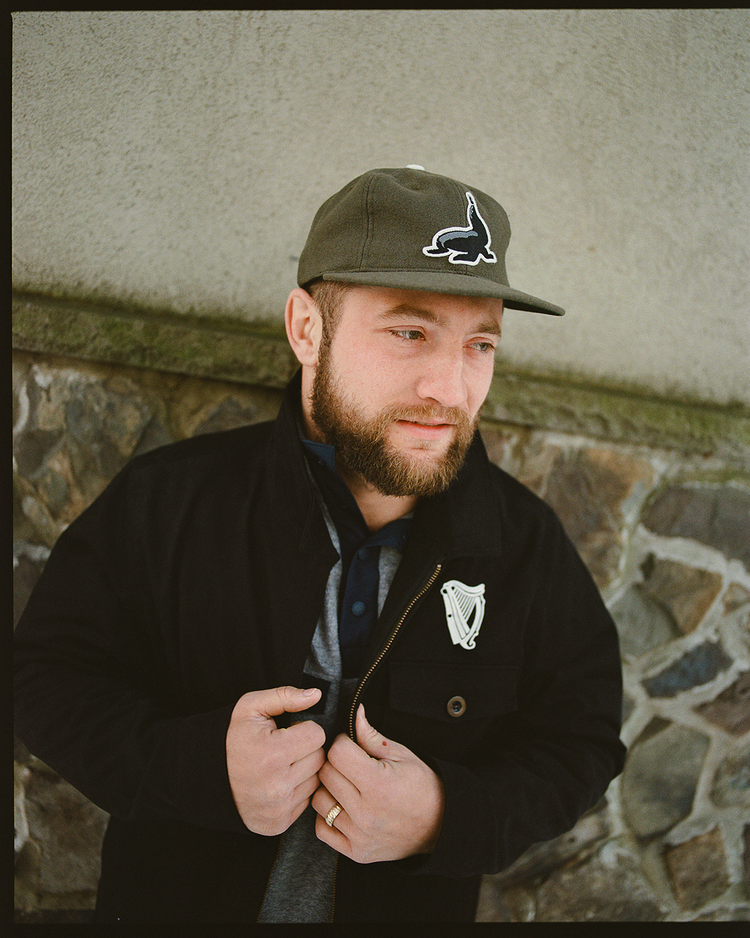 A bearded man outdoors by a stone wall wears an Ebbets Field Guinness Seal Authentic Wool Ballcap and a dark jacket, looking slightly to the side with a calm expression as he adjusts his jacket with both hands.