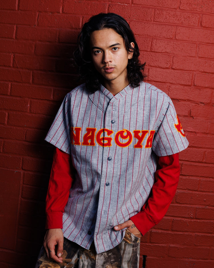 A person with long dark hair wears the Ebbets Field Nagoya Club 1938 Authentic Wool Road Jersey, featuring red NAGOYA lettering, while standing against a red brick wall.