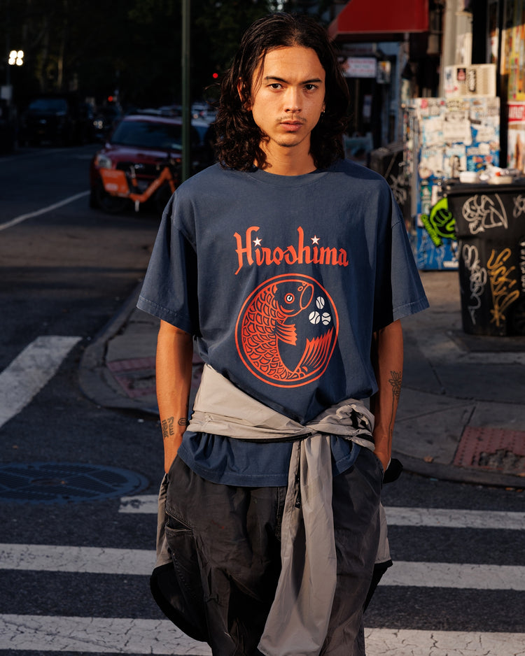 A person with long dark hair stands at a city crosswalk in an Ebbets Field Hiroshima Carp Pigment-Dyed Short Sleeve Graphic T-Shirt, jacket tied at the waist, hands in pockets, with storefronts and street signs behind.