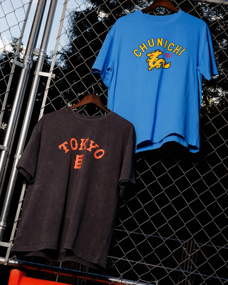 Two T-shirts hang on a fence: a blue Chunichi Dragons Short Sleeve Graphic T-Shirt by Ebbets Field with a yellow dragon, and a black vintage tee with red TOKYO lettering. Trees and sky fill the background.