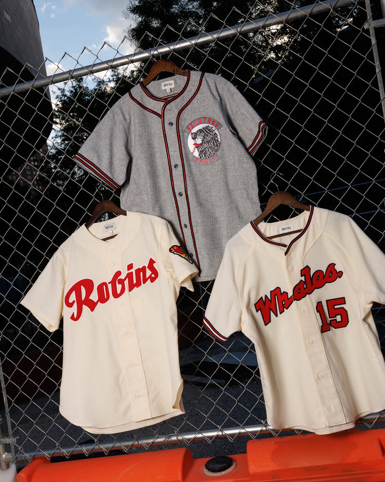 Three vintage-style jerseys, including the Ebbets Field Tokio Senators 1936 Authentic Wool Road Jersey, hang on a chain-link fence with trees and sky in the background.