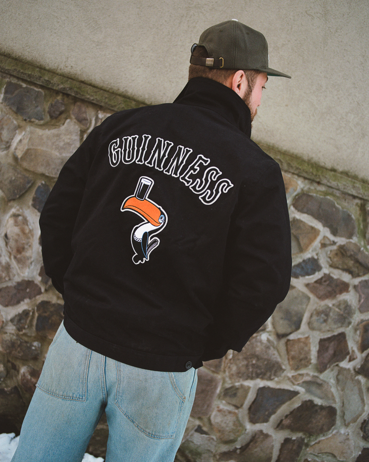Someone wearing the Guinness Cotton Grounds Crew Jacket by Ebbets Field, featuring a colorful toucan graphic on the back, stands with their back to the camera in front of a stone wall, paired with light blue jeans and a green cap.