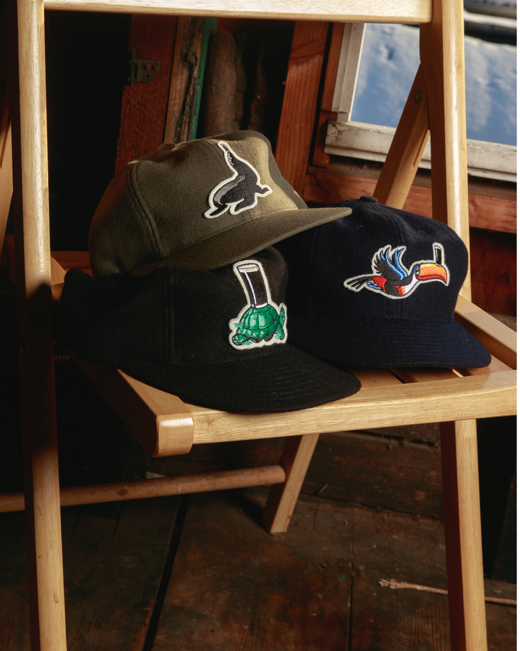Three baseball caps sit on a wooden chair: an olive green Ebbets Field hat with a black rabbit, a black cap with a tortoise and golf flag, and the Ebbets Field Guinness Tortoise Authentic Wool Ballcap featuring a colorful bird patch.
