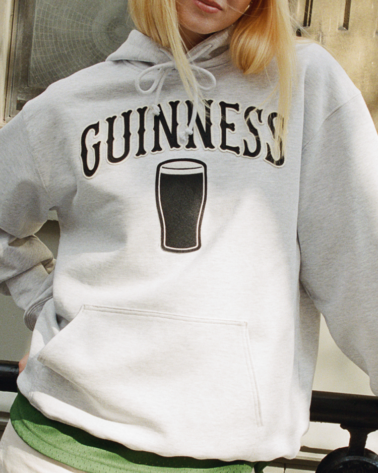 A person with long blond hair partially out of frame wears the Ebbets Field Guinness Heavyweight Cotton Terry Pullover Hoodie, featuring 