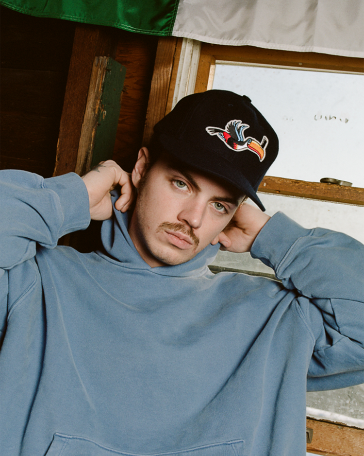 A young man in a blue hoodie wears an Ebbets Field Guinness Flying Toucan Authentic Wool Ballcap, standing indoors with his hands behind his neck. A partially visible flag hangs above him as he looks at the camera.