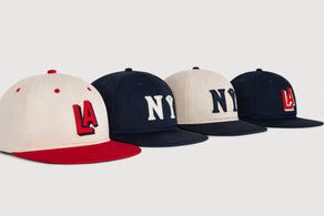 Four baseball caps are displayed in a row on a white background. Two caps are beige with red or navy brims and LA or NY logos, and two are navy with NY or LA logos in white or red.