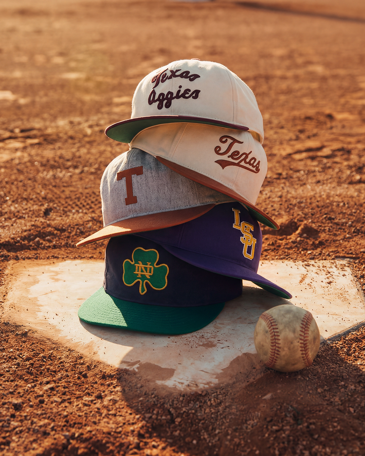 An Ebbets Field University of Texas 1943 Cotton Twill Ballcap sits atop a stack of college hats on a pitcher’s mound, next to a worn baseball, on a sunlit dirt field.