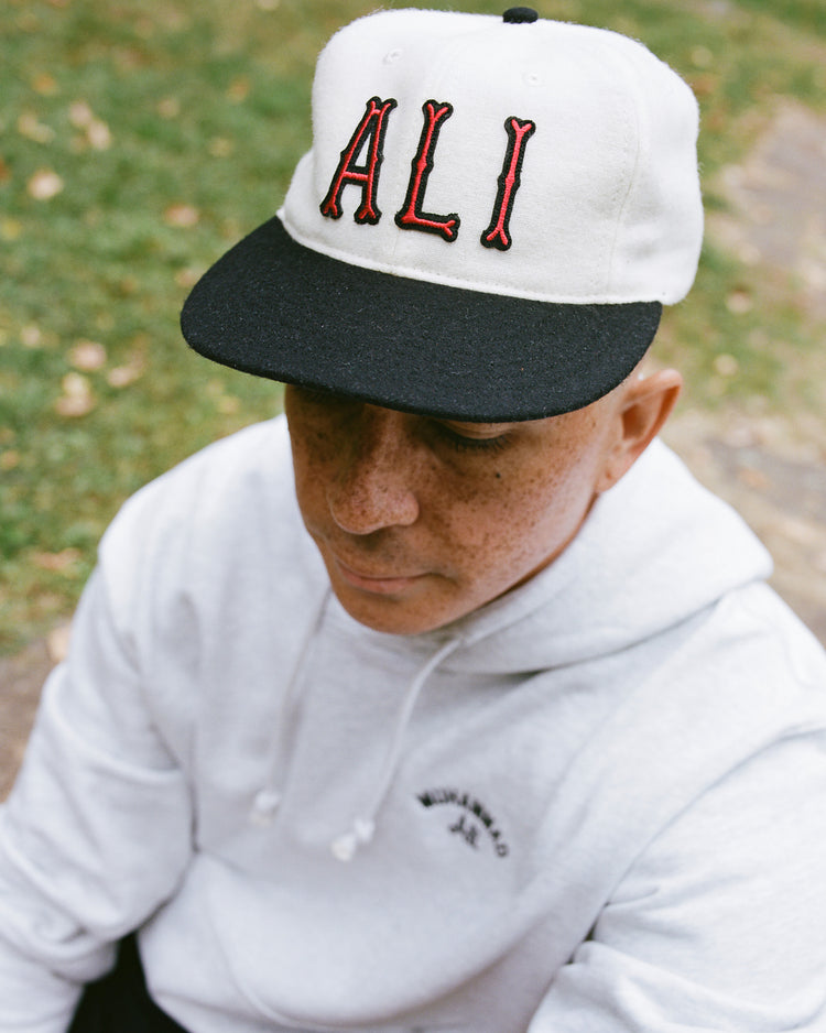 A person wears the Muhammad Ali Vintage Inspired Wool Ballcap by Ebbets Field, sitting on a stone path outdoors. Grass and scattered leaves evoke the calm after the Thrilla in Manila, channeling Ali's enduring legacy.
