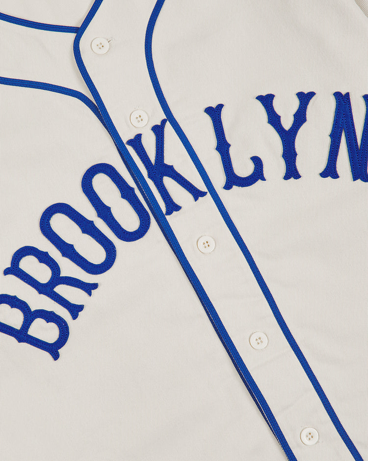 Introducing the Brooklyn Royal Giants 1919 Cotton Twill Baseball Jersey by Ebbets Field—a white cotton twill jersey with blue piping and bold blue “BROOKLYN” lettering across the front, inspired by the classic 1919 uniform worn by the Brooklyn Royal Giants. The front features traditional white buttons running down the center.