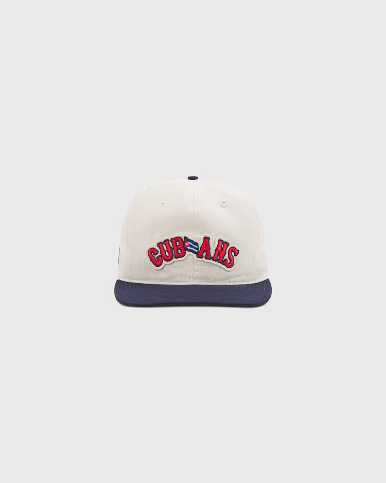 The Ebbets Field New York Cubans Authentic Cotton Ballcap features cream-colored cotton twill with a navy blue brim. The front displays “CUBANS” embroidered in red with a blue outline, honoring the historic New York Cubans from the Negro League. The background is plain and light gray.