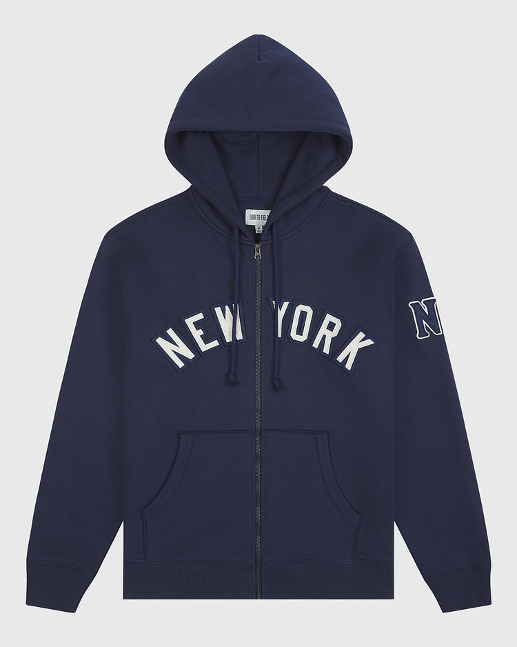 The Ebbets Field Black Yankees Heavyweight Cotton Terry Full-Zip Hoodie features a navy blue design with “NEW YORK” in white block letters across the chest, an “N” patch on the left sleeve, front pockets, and a drawstring hood—drawing inspiration from classic Negro League apparel.