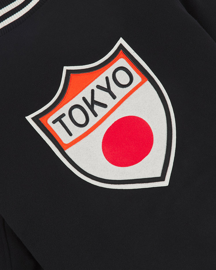 A close-up of the Ebbets Field Tokyo Giants All-Wool Varsity Jacket showcases a black fabric adorned with a shield-shaped patch. The patch features the word “TOKYO” above a red circle, reminiscent of the Japanese flag, and is inspired by the Tokyo Giants and Japanese professional baseball. Accented in black, white, and orange, it’s perfectly suited to this premium all-wool varsity jacket.