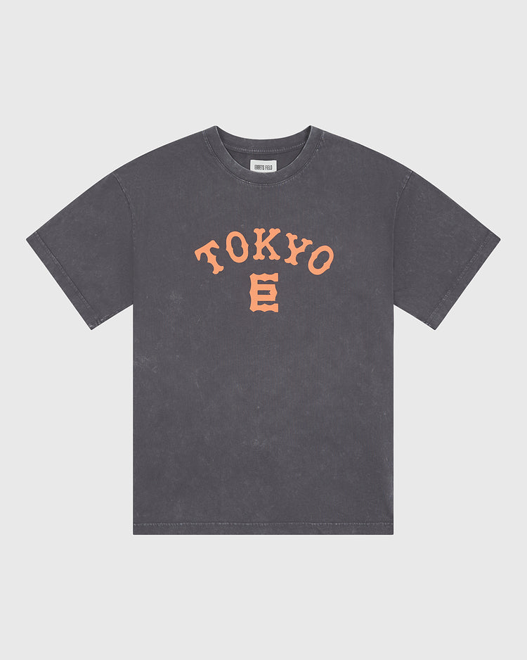 The Ebbets Field Tokyo Giants Pigment-Dyed Short Sleeve Graphic T-Shirt features a gray, pigment-dyed finish and displays bold orange “TOKYO” lettering alongside a stylized E graphic on the front, all inspired by the Tokyo Giants. The shirt is presented against a plain white background.
