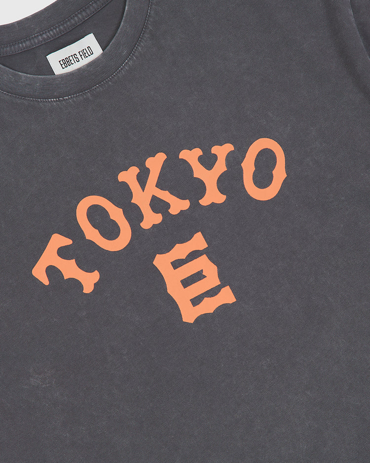 The Ebbets Field Tokyo Giants Pigment-Dyed Short Sleeve Graphic T-Shirt is a black tee featuring bold orange arched “TOKYO” text on the front with a stylized “E” below, subtly evoking the spirit of the Tokyo Giants. The inner label is marked with EBBETS FIELD.