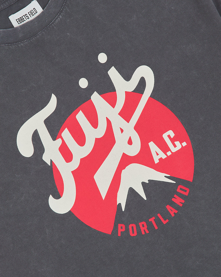 The Fuji Athletic Club Pigment-Dyed Short Sleeve Graphic T-Shirt by Ebbets Field features a dark gray, pigment-dyed fabric with a bold graphic design: “Fuji A.C. PORTLAND” printed in white and red letters over a striking red circle, showcasing a mountain graphic that pays tribute to Japanese American baseball heritage. The shirt includes an Ebbets Field-branded neck tag for added authenticity.