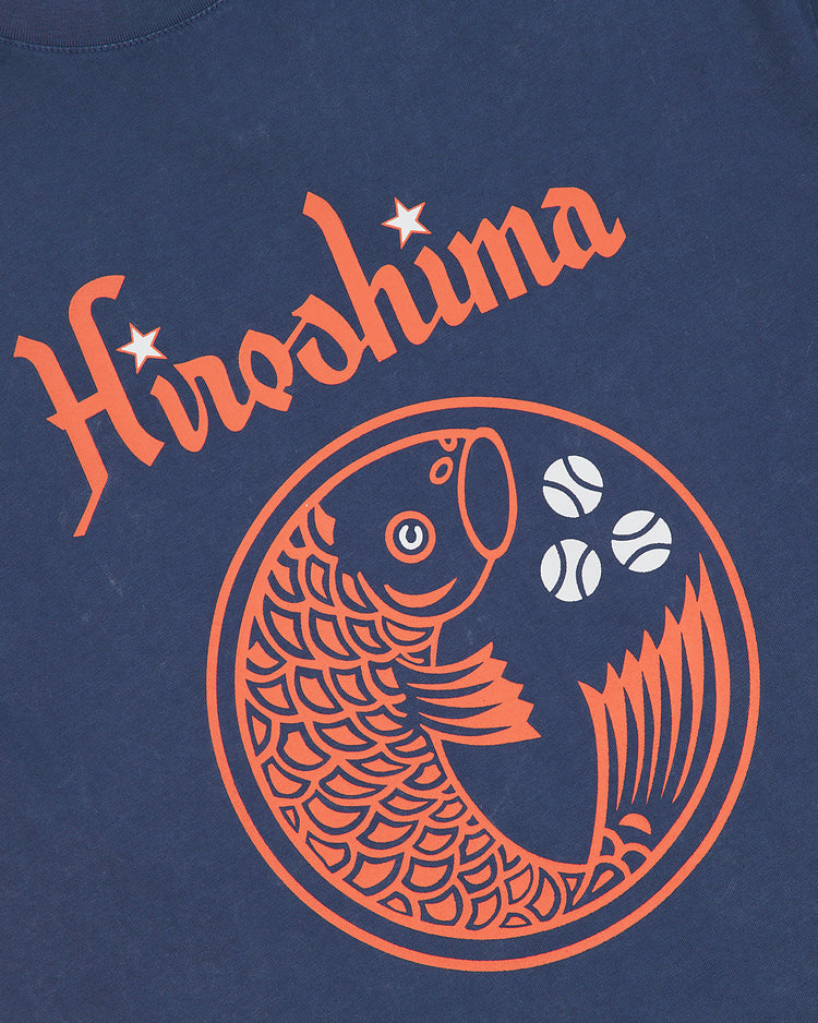 The Ebbets Field Hiroshima Carp Pigment-Dyed Short Sleeve Graphic T-Shirt features a navy blue color with “Hiroshima” in orange stylized letters above an orange fish and three baseballs.