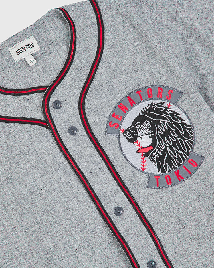 The Ebbets Field Tokio Senators 1936 Authentic Wool Road Jersey is a gray button-up featuring black and red piping, a bold embroidered bear patch, and “Senators Tokio” lettering on the chest. Inspired by the classic 1936 Tokio Senators, this vintage baseball jersey captures timeless style, with an Ebbets Field label on the inside.