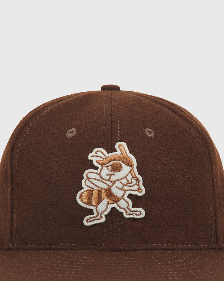 The Ebbets Field Salt Lake City Bees 1959 Wool Ballcap is a brown wool cap featuring an embroidered cartoon bee holding a baseball bat on the front, inspired by the 1959 Salt Lake City Bees of the Pacific Coast League.