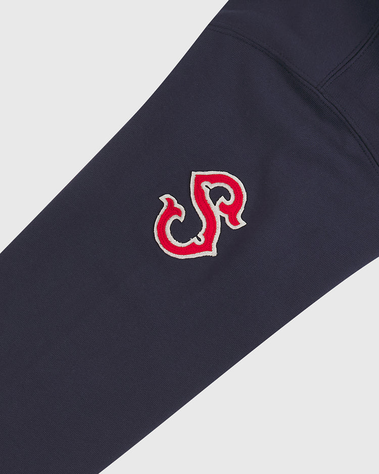 A close-up of a navy blue fabric sleeve showcases a red and white embroidered logo with the stylized letters J and P intertwined, evoking classic details from an authentic 1941 Seattle Rainiers Heavyweight Cotton Terry Pullover Hoodie by Ebbets Field.