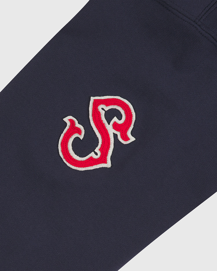 Close-up of a dark navy heavyweight cotton terry fabric featuring a stitched red and white embroidered emblem. The emblem showcases a stylized, swirling design that blends letters, reminiscent of a vintage collegiate or athletic varsity logo—perfectly capturing the classic charm of the Ebbets Field Seattle Rainiers 1941 Heavyweight Cotton Terry Pullover Hoodie.