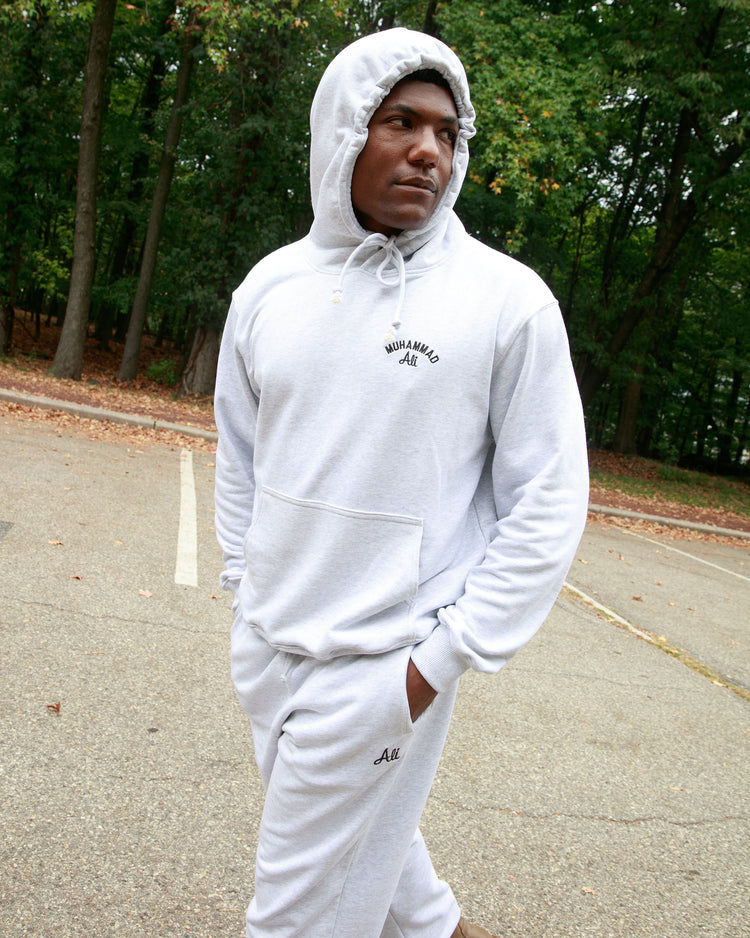 A man stands in a tree-lined parking lot wearing an Ebbets Field Muhammad Ali Chest Embroidered Heavyweight Cotton Terry Pullover Hoodie, hood up and hands in his pockets, channeling classic boxing training style.