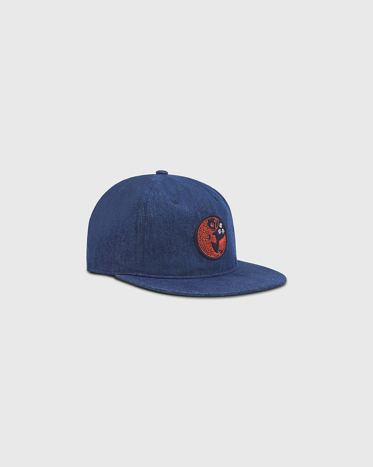 The Ebbets Field Hiroshima Carp Indigo Cotton Denim Twill Ballcap, inspired by Japanese baseball, features a flat brim and a red and black embroidered circular Hiroshima Carp patch on the front. The blue denim cap is displayed on a light gray background.