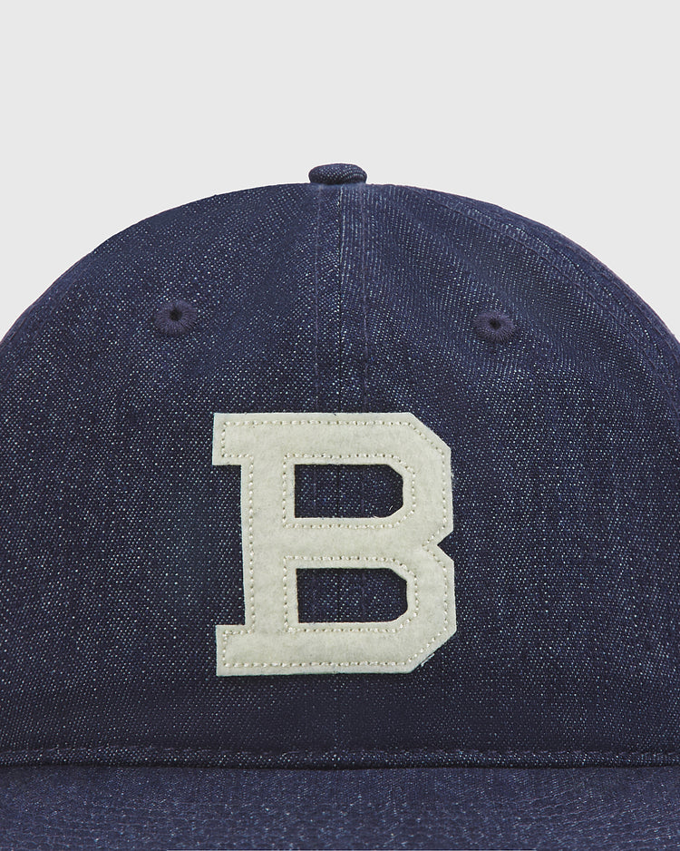 A close-up of the Ebbets Field Brooklyn Bushwicks Indigo Cotton Denim Ballcap, featuring a prominent white letter B patch on the front, evokes the look of a vintage baseball cap against a plain light background.
