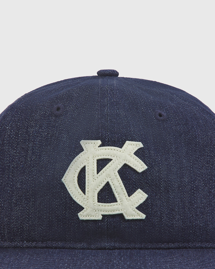 The Ebbets Field Kansas City Monarchs Indigo Cotton Denim Ballcap is a dark blue denim baseball cap with a white embroidered KC logo on the front, inspired by the legacy of the Kansas City Monarchs and styled after a vintage ballcap, displayed against a plain white background.