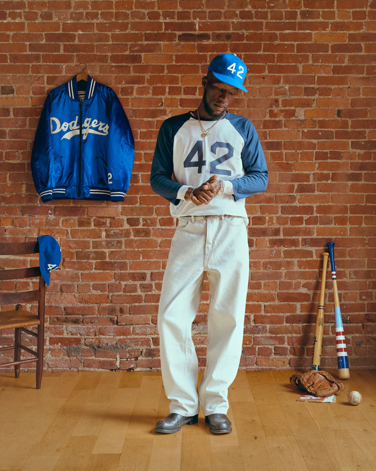Wearing the Ebbets Field Jackie Robinson 42 Vintage Baseball Tee and a blue 42 cap, a man stands before a brick wall filled with Dodgers gear, adjusting his sleeve as hes surrounded by classic memorabilia and vintage baseball shirts.