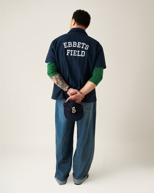 A person stands facing away, hands behind their back holding a navy cap with a white logo. They wear a navy shirt reading EBBETS FIELD, a green long-sleeve top underneath, and loose blue jeans.