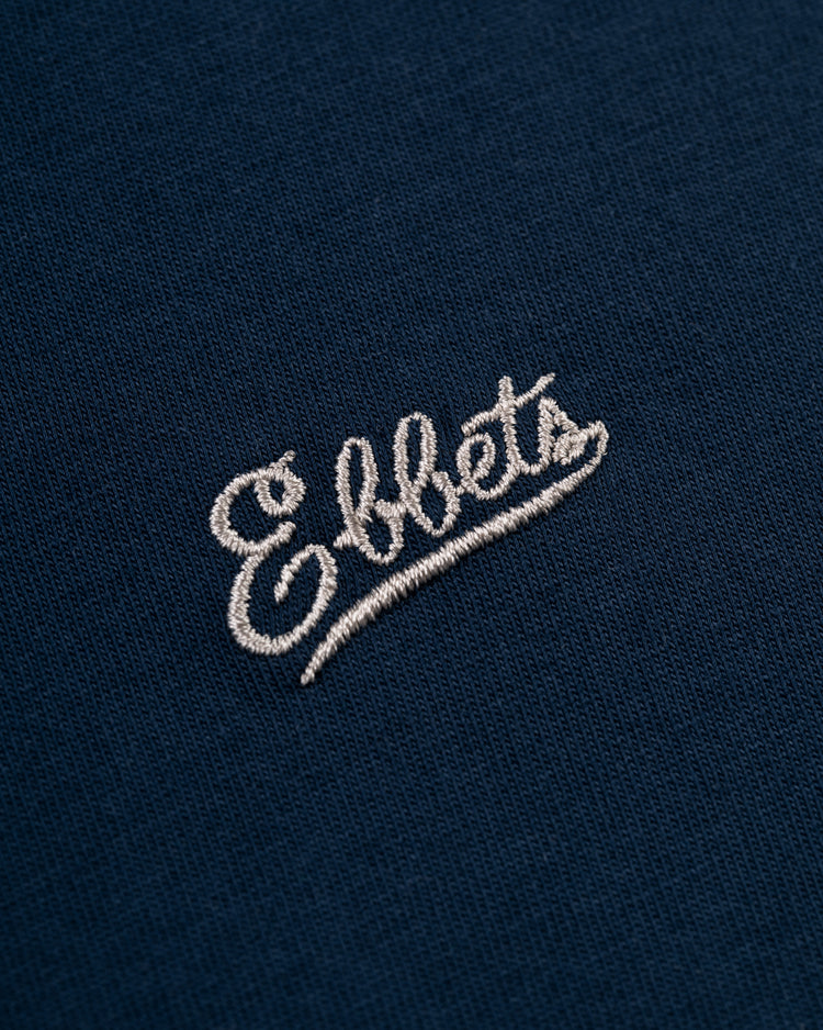 Close-up of the white embroidered Ebbets Field Flannels logo on dark blue fabric of the Ebbets Chest Logo Short Sleeve Tee—a true wardrobe staple.